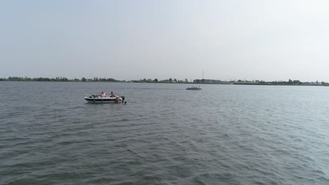 Drohnen-Slomo-Eines-Kleinen-Bootes-Auf-Dem-Fluss-Mit-Männlichen-Freunden,-Die-Schwimmen-Und-Am-Boot-Hängen,-Mit-Vorbeifahrendem-Boot-Im-Hintergrund