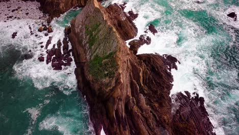 Jagged-Rock-Cliffs-in-Shimmering-Turquoise-Ocean,-Rising-Drone-Reveal