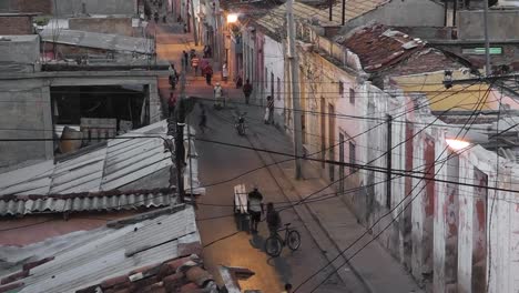 Vista-De-Pájaro-Sobre-La-Típica-Vida-Callejera-Cubana-Al-Atardecer-Con-Luces-De-Calle-Suaves,-Cables-De-Electricidad-Y-Teléfono-Colgando,-Niños-Jugando,-Adultos-Caminando-Y-Motocicletas-Pasando