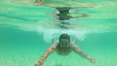Man-diving-into-shallow-blue-sea-water-and-swimming-underwater-towards-camera-smiling---showing-thumbs-up
