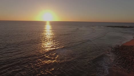 Sunrise-drone-shot-over-water-and-beach-with-sun-at-center-of-frame