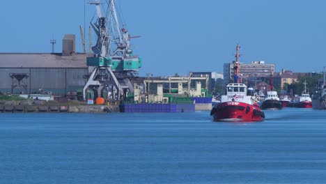 Remolcador-De-Puerto-Saliendo-Del-Puerto-De-Liepaja-En-Un-Día-Caluroso-Y-Soleado,-Grúas-Portuarias-En-El-Fondo,-Toma-Amplia-Desde-La-Distancia