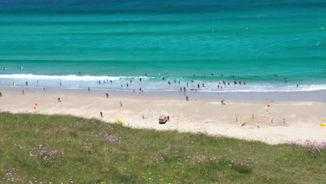 Ocean-waves-along-the-wide-sandy-shore-at-Sennen-Cove-in-Cornwall-with-surfers-and-tourists-walking-on-the-beach