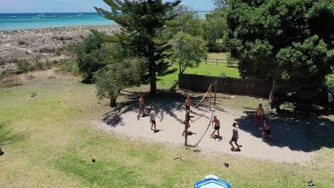 Vista-Aérea-De-Personas-Jugando-Voleibol-En-La-Arena-Bajo-Las-Sombras-De-Los-árboles-Junto-Al-Mar-En-Un-Día-Soleado