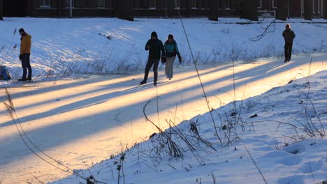 Pan-Zeigt-Eislaufende-Menschen-Auf-Zugefrorenen-Wasserstraßen-Mit-Schneebedeckten-Ufern-Des-Flusses-Berkel-In-Blautönen-Und-Silhouetten-Von-Menschen,-Die-In-Den-Sonnenstrahlen-Des-Späten-Nachmittags-Lange-Schatten-Werfen