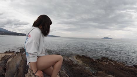 A-brunette-woman-in-20s-sitting-alone-on-a-seashore-rock-along-a-coast-in-Greece-on-a-partly-sunny-day