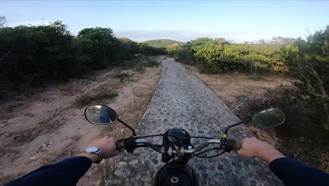 Motorcyclist-rides-through-coastal-rocks-and-shrubs,-arrives-to-ocean,-pov