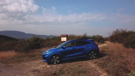 Aerial-top-view-over-a-blue-Ford-Puma-parked-next-to-a-country-crossroads,-on-top-of-a-hill