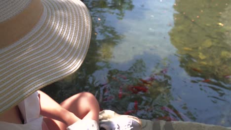 Top-down-view-of-girl-in-summer-hat-giving-food-to-Koi-fish-in-pond-with-reflections