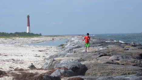 Barnegat,-Nueva-Jersey---14-De-Agosto-De-2020:-Un-Niño-Caminando-En-El-Embarcadero-De-Roca-En-El-Embarcadero-De-Barnegat-Con-El-Faro-Al-Fondo