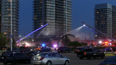 Escaleras-Aéreas-De-Camiones-De-Bomberos-Rociando-Agua-En-El-Perímetro-Del-Incendio-Con-Tráfico-Rodado-En-Primer-Plano