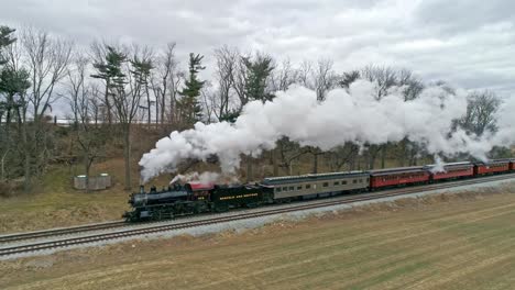 Eine-Drohnenansicht-Einer-Dampflokomotive-Mit-Personenwagen,-Die-Sich-An-Einem-Wintertag-Mit-Voller-Dampfkraft-über-Die-Landschaft-Nähern