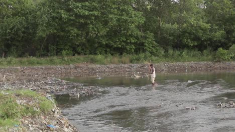 A-Male-Villager-Walks-In-A-River-With-A-Stick,-Garbage-Waste-Site-At-Slums-In-Pakistan