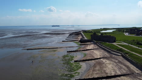 Vuelo-De-Drones-Sobre-Marismas-Del-Estuario-Y-Una-Playa-Separada-Por-Viejos-Espigones-De-Madera-Durante-La-Marea-Baja,-Con-Un-Malecón-Y-Una-Guarnición-Militar-Abandonada-En-El-Lado-Derecho,-En-Un-Día-Claro-Y-Caluroso-De-Verano