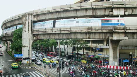 BTS-Sky-Train-on-rail-tracks-over-traffic-through-busy-intersections-at-rush-hour-in-Bangkok-which-is-controlled-by-traffic-lights,-there-is-heavy-traffic-every-day