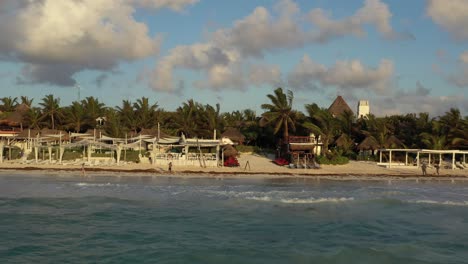 Aerial-backwards-shot-of-person-cleaning-water-plants-of-sandy-beach-in-front-of-outdoor-bar-in-Tulum,Mexico