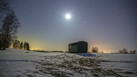 Stars-moving-in-sky-during-clear-night-above-tiny-cabin-in-snowy-landscape,-timelapse