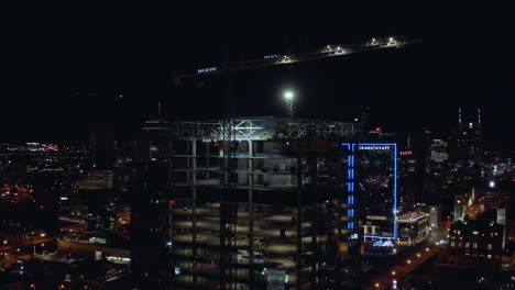 Ascending-night-aerial-up-tower-under-construction-with-cranes-in-city,-4K