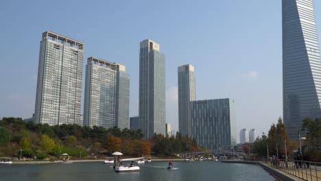 Parque-Central-De-Songdo-En-Incheon,-Corea-Del-Sur,-Con-Personas-Que-Disfrutan-De-Viajar-En-Botes-Y-Caminar-Junto-Al-Lago-Con-Rascacielos-De-Gran-Altura-Contra-El-Cielo-Despejado,-Torre-Posco---Establecimiento-De-Tiro