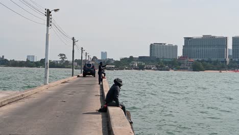 A-man-sitting-then-fixes-its-rod-before-casting-while-other-already-reeling-in-as-a-truck-passes-by,-Pattaya-Fishing-Dock,-Chonburi,-Thailand