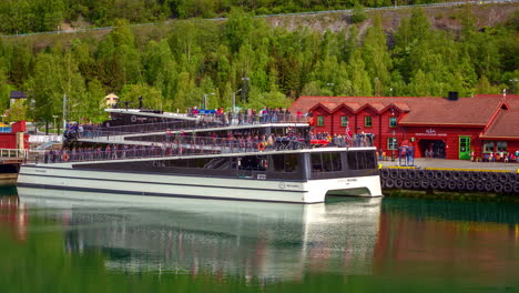 Ferry-Catamarán-Eléctrico-Con-Pasajeros-Saliendo-Del-Puerto-En-Aurlandsfjord-En-Flam,-Aurland,-Noruega-Con-El-Centro-De-Visitantes-De-Flam-En-Segundo-Plano