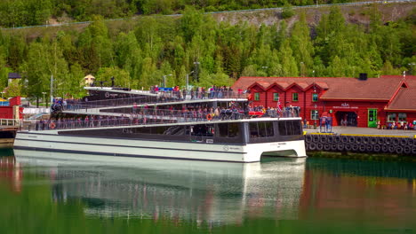 Elektrische-Katamaran-Fähre-Mit-Passagieren,-Die-Den-Hafen-Am-Aurlandsfjord-In-Flam,-Aurland,-Norwegen-Mit-Flam-Besucherzentrum-Im-Hintergrund-Verlassen