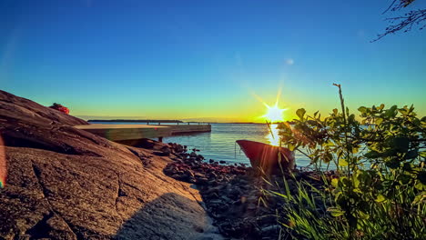 Hyperlapse-shot-of-peaceful-rocky-shoreline-with-sea-and-jetty-during-golden-sunset-at-horizon---Ancient-boat-anchored-at-rivershore
