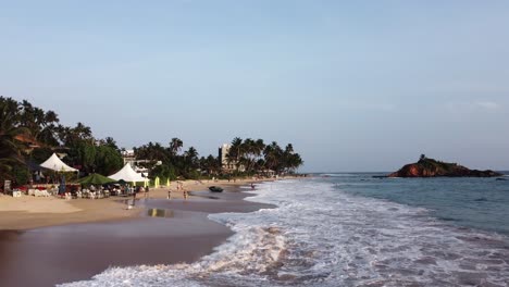 Aerial-dolly-in-of-people-relaxing-in-sand-shore-near-sea-waves-and-palm-tree-rainforest,-Mirissa-Beach,-Sri-Lanka