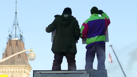 Dos-Hombres-De-Pie-Y-Portando-Pancartas-Levantando-La-Mano-Durante-La-Protesta-Del-Convoy-De-La-Libertad