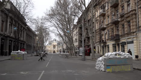 People-walk-past-concrete-barricades,-one-of-which-is-sprayed-painted-in-yellow-and-blue-spray-paint-with-the-words,-“Liberté,-égalité,-fraternité”-in-front-of-the-Odesa-Opera-and-Ballet-theatre