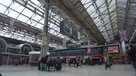 Vista-Interior-De-La-Estación-Victoria-En-Londres-Con-Bandera-Británica-Colgando-Del-Techo-Y-Pasajeros-Caminando