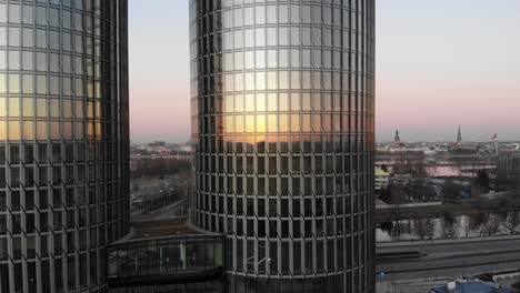 Aerial-view-of-two-glass-skyscrapers-on-golden-sunset-with-Riga-cityscape-in-the-background-in-Latvia