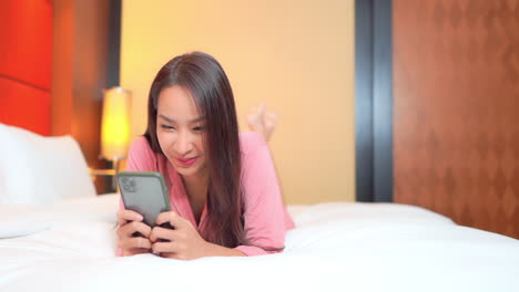 Close-up-of-a-young-woman-lying-on-her-stomach-across-the-bed-as-she-texts-and-reads-messages-from-a-smartphone