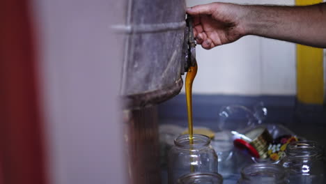 Farmer-Beekeeper-Filling-Glass-Jar-with-Freshly-Extracted-Golden-Bee-Honey,-Flowing-Dripping-then-Turns-off-the-Handle-of-the-Extractor-Faucet-with-his-Hand,-Traditional-Honey-Production