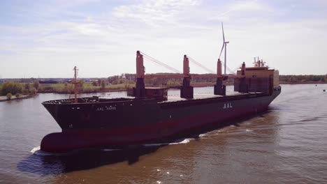 Aerial-Port-Side-View-Of-Aal-Paris-Cargo-Ship-Travelling-Along-Oude-Maas-With-Still-Wind-Turbine-In-Background