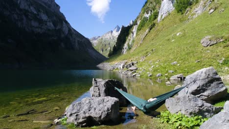 guy-relaxing-in-a-hammock-outdoor-at-a-mountain-lake-in-the-swiss-alps,-aerial-flying-over