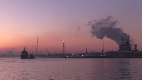 a-cargo-ship,-assisted-by-a-tugboat-coming-in-at-dawn-at-the-port-of-Antwerp-and-its-industry