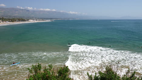 Eine-Person,-Die-An-Einem-Sonnigen-Strandtag-Auf-Einer-Blauen-Ozeanwelle-Mit-Der-Stadt-Santa-Barbara,-Kalifornien,-Im-Hintergrund-In-Zeitlupe-Surft