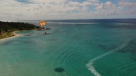 Aerial-shot-of-a-man-practicing-parasailing-over-the-coast,-with-his-shadow-on-the-water,-beside-a-tree-covered-beach