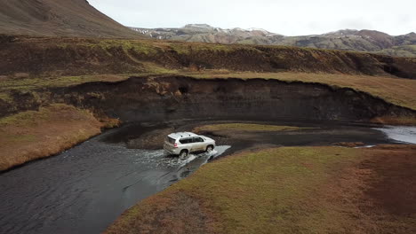 Cuatro-Ruedas-En-Todoterreno-Y-Río-Poco-Profundo-En-El-Campo-De-Islandia-Bajo-Colinas-Volcánicas,-Vista-Aérea