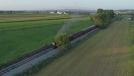 Vista-Aérea-De-Un-Motor-De-Vapor-De-1924-Con-Un-Tren-De-Pasajeros-Que-Viaja-A-Lo-Largo-De-La-Campiña-Amish-Mientras-Las-Puestas-De-Sol-En-Un-Día-De-Verano-Visto-Por-Un-Dron