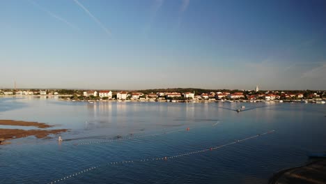 Panorama-Aéreo-De-Una-Ciudad-Nin,-Playa-De-Arena-Croata-Y-Niños-Jugando,-Con-Laguna-Y-Barco-Acercándose-Al-Atardecer