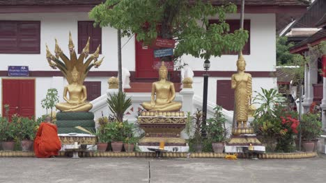 Mönch-Mit-Orangefarbener-Robe-Betet-Im-Buddhistischen-Tempel-Mit-Drei-Goldenen-Buddha-Statuen-In-Laos,-Luang-Prapang
