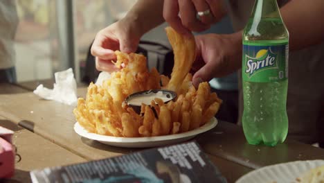 Manos-Agarrando-Un-Plato-De-Cebolla-Floreciente-Frita-Con-Salsa-En-La-Mesa-Y-Una-Botella-De-Refresco-Al-Lado