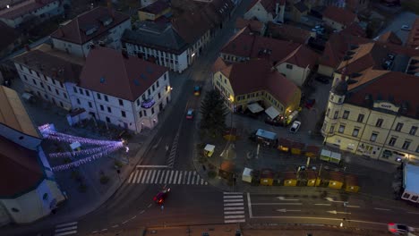 Las-Luces-De-Navidad-Se-Encienden-En-La-Plaza-De-La-Pequeña-Ciudad,-El-Tiempo-Aéreo-Del-Mercado-De-Navidad-Del-Anochecer-A-La-Noche,-Los-Autos-Pasan-Por-La-Calle,-Tiro-Constante