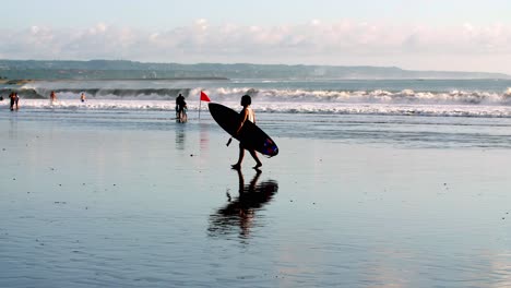 Surfer-walking-along-the-beach-carrying-board-in-slow-motion,-red-flag-on-sand-to-warn-of-high-surf-and-currents,-handheld-shot