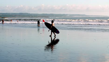 Surfista-Caminando-Por-La-Playa-Con-Tabla-En-Cámara-Lenta,-Bandera-Roja-En-La-Arena-Para-Advertir-Sobre-Olas-Y-Corrientes-Altas,-Tiro-De-Mano