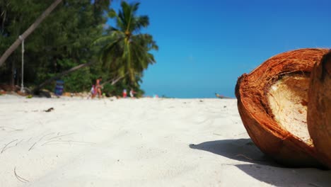 Nahaufnahme-Der-Geöffneten-Geknackten-Kokosnuss-Auf-Dem-Weißen-Sand-Mit-Bikini-Mädchen,-Die-Im-Hintergrund-Am-Strand-Entlang-Spazieren