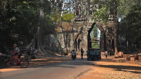 Medium-Exterior-Slow-Motion-Shot-of-Motorcycle-Coming-Through-Ancient-Gate-and-Tourists-in-the-Background-Walking-by-a-Huge-Ancient-Tree-With-Roots-in-the-of-Temple-Area