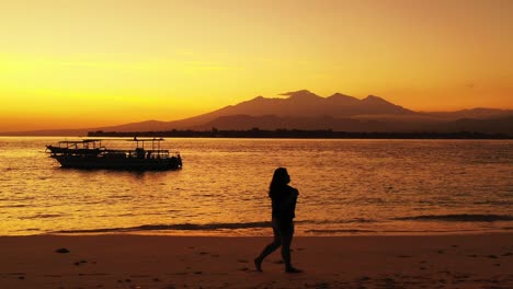 Silhouette-Eines-Mädchens,-Das-Bei-Sonnenuntergang-Am-Ruhigen-Exotischen-Strand-Spazieren-Geht,-Mit-Goldenem-Himmel,-Der-Sich-Auf-Einer-Ruhigen-Lagune-Voller-Boote-In-Bali-Widerspiegelt